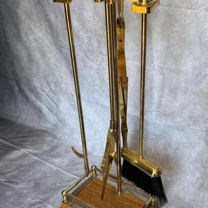 Puede incluir: Un juego de herramientas para chimenea de latón con mangos de madera y un fuelle a juego. El juego incluye un atizador, pinzas, pala y cepillo, todo exhibido en un soporte de madera y latón. El fuelle es marrón con un emblema decorativo de latón.