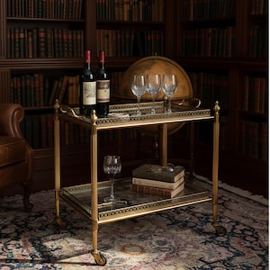 May include: A gold drinks trolley with two glass shelves, displaying wine bottles, wine glasses, and books. The cart features a decorative filigree design and is set in a library with a globe.