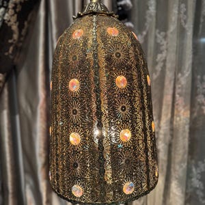 May include: An ornate, bell-shaped lamp with a dark bronze finish. The lamp shade has a detailed, openwork design with circular patterns and is decorated with iridescent, circular embellishments. The lamp's base and top finial have decorative details.
