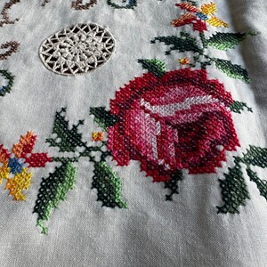 May include: Close-up of a white linen tablecloth with a red rose and green leaves embroidered in cross-stitch. A small white crocheted circle is in the upper left corner.
