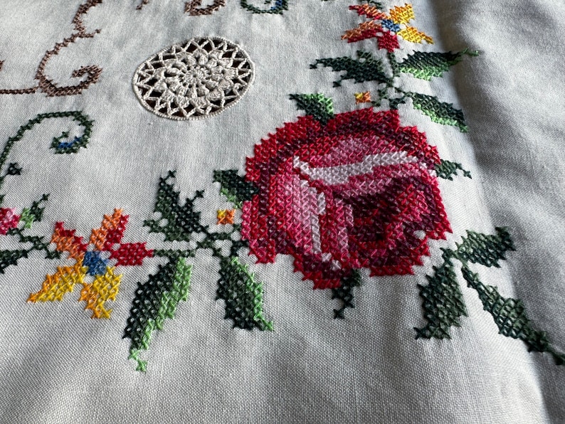 May include: Close-up of a white linen tablecloth with a red rose and green leaves embroidered in cross-stitch. A small white crocheted circle is in the upper left corner.