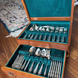 Peut inclure: Un coffret à couverts en bois à deux tiroirs, doublé de velours turquoise. Les tiroirs sont remplis de couverts en argent ornés, notamment des cuillères, des fourchettes et des couteaux. Le coffret est ouvert, affichant le contenu. Le bois a une teinte brun-rougeâtre chaude.
