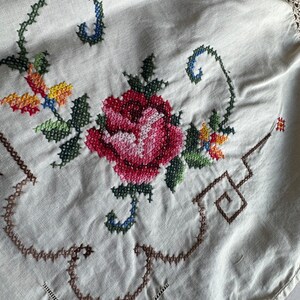 May include: A white linen tablecloth with a hand-stitched floral design. The design features a red rose with green leaves and stems, and smaller yellow and orange flowers. The tablecloth has a crocheted border.
