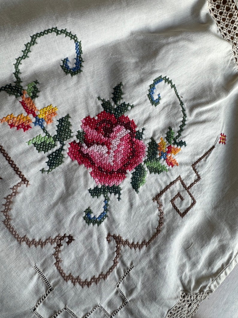 May include: A white linen tablecloth with a hand-stitched floral design. The design features a red rose with green leaves and stems, and smaller yellow and orange flowers. The tablecloth has a crocheted border.
