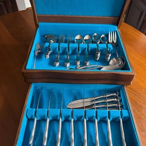 May include: A wooden box with a blue velvet interior, containing a set of cutlery. The cutlery includes forks, knives, and spoons, all arranged neatly in the box. The box has a hinged lid and a latch.