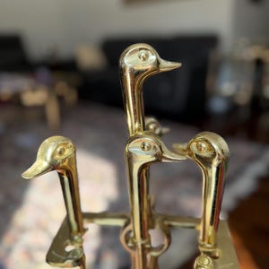 Puede incluir: Un juego de herramientas de chimenea de latón con mangos de cabeza de pato. Las herramientas incluyen un atizador, una pala y un cepillo, todos con un acabado dorado brillante. Las cabezas de pato están detalladas.