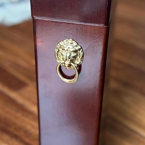 Puede incluir: Una caja de madera rectangular y alta con un acabado marrón oscuro. Una cabeza de león dorada con un asa de anillo está fijada en la parte delantera. La caja parece ser un artículo de almacenamiento decorativo.