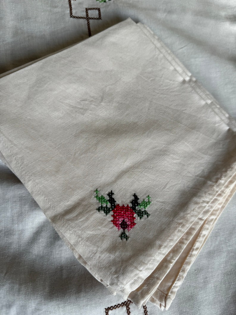May include: A white linen napkin with a red and green embroidered flower design.