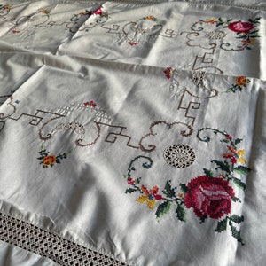 May include: White linen tablecloth with delicate embroidered floral designs in shades of red, green, yellow, and brown. The tablecloth features a crocheted border.