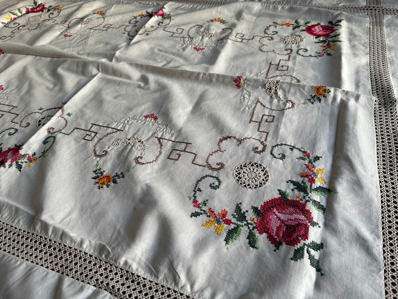 May include: White linen tablecloth with delicate embroidered floral designs in shades of red, green, yellow, and brown. The tablecloth features a crocheted border.