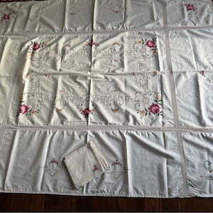 May include: A white tablecloth with a delicate crocheted border and embroidered floral designs. The tablecloth is made of a lightweight fabric and features a repeating pattern of roses and leaves.