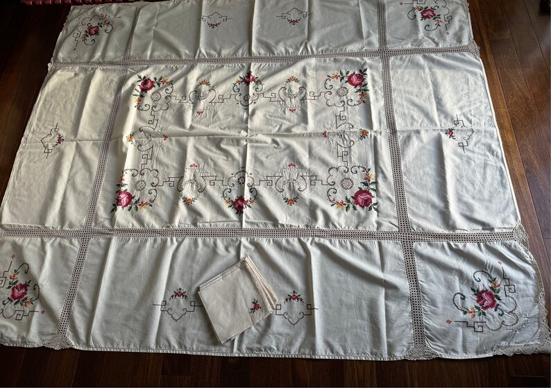 May include: A white tablecloth with a delicate crocheted border and embroidered floral designs. The tablecloth is made of a lightweight fabric and features a repeating pattern of roses and leaves.
