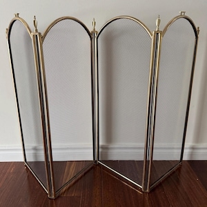 Puede incluir: Una pantalla de chimenea de latón y negra con un diseño de cuatro paneles. Cada panel tiene una fina pantalla de malla enmarcada por metal color latón. La parte superior de cada panel tiene forma de arco y la pantalla está decorada con pequeños pináculos.