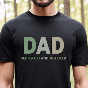 May include: A black t-shirt with a green, white, and gray gradient "DAD" graphic and the text "DEDICATED AND DEVOTED" underneath.