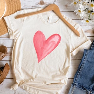 May include: A cream colored t-shirt with a pink watercolor heart design. The shirt is on a hanger and there are jeans, sandals, and a straw hat in the background.