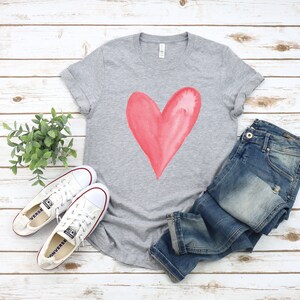 May include: A gray t-shirt with a large, pink watercolor heart design. The shirt is laid out on a white wooden surface with a pair of white canvas shoes and blue jeans.