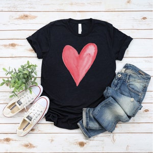 May include: A black t-shirt with a red watercolor heart design. The shirt is paired with blue jeans and white sneakers.
