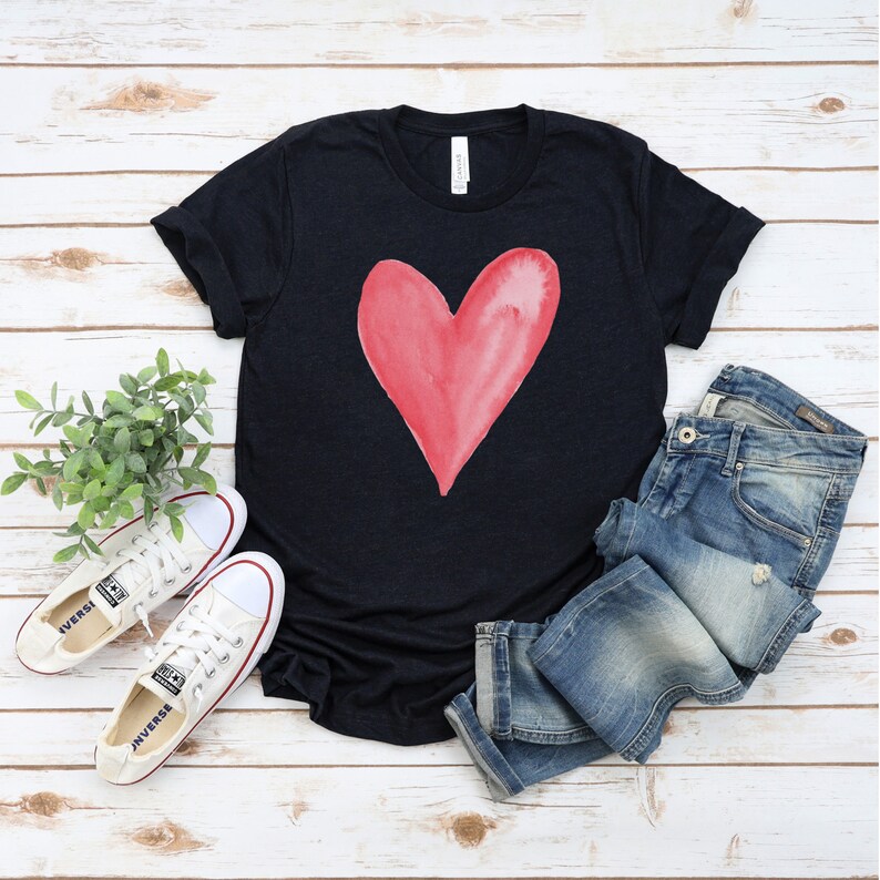 May include: A black t-shirt with a red watercolor heart design. The shirt is paired with blue jeans and white sneakers.