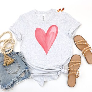 May include: A heather grey t-shirt with a large, pink watercolor heart design. The shirt is shown with denim shorts and brown sandals.