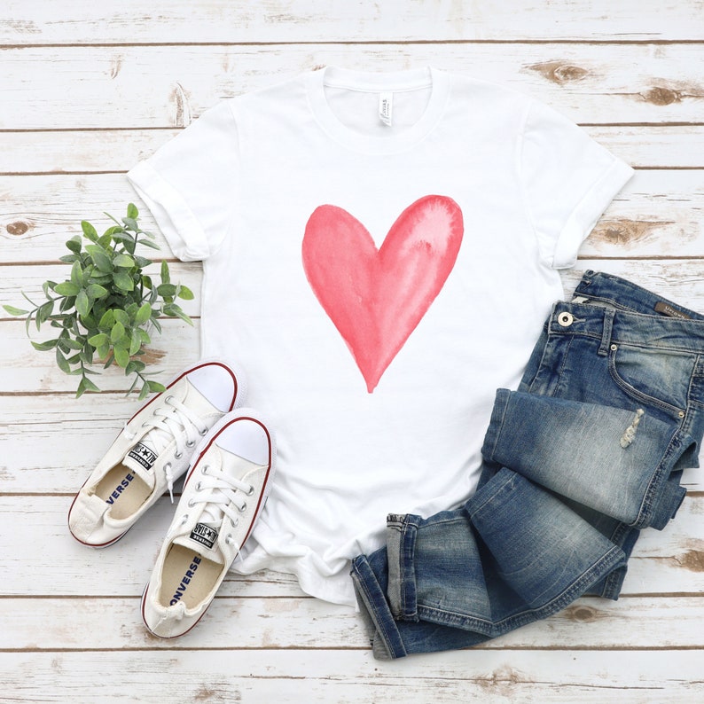 May include: White t-shirt with a pink watercolor heart design. The shirt is paired with blue jeans and white sneakers.