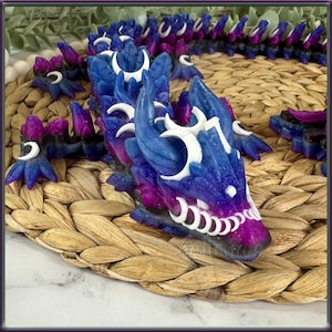 Pode incluir: Uma figura de dragão azul e roxo com detalhes brancos e um acabamento brilhante. O dragão está enroscado com a cabeça apoiada na cauda. A figura é feita de resina e mede aproximadamente 25 cm de comprimento.