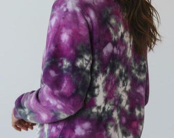 May include: A tie-dye sweatshirt in shades of purple, gray, and white. The long-sleeved top has a crew neck and a relaxed fit. The fabric has a swirling pattern, creating a unique and artistic look. The sweatshirt is paired with blue jeans.
