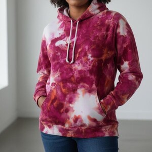 May include: A tie-dye hoodie in shades of pink, white, and orange. The hooded sweatshirt has a kangaroo pocket and white drawstring. The model is wearing blue jeans. The hoodie is a casual, comfortable garment.