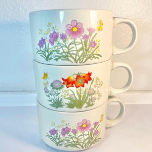 May include: Three white ceramic mugs with floral designs. The mugs are stacked on top of each other. The top mug has a pink and purple flower design, the middle mug has an orange flower design, and the bottom mug has a pink and purple flower design.