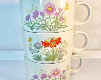May include: Three white ceramic mugs with floral designs. The mugs are stacked on top of each other. The top mug has a pink and purple flower design, the middle mug has an orange flower design, and the bottom mug has a pink and purple flower design.