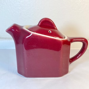 May include: A burgundy ceramic teapot with a lid and a small spout. The teapot has a geometric shape and a glossy finish.