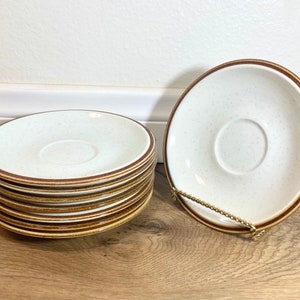 Pode incluir: Uma pilha de seis pires de cerâmica marrom e branco salpicados com uma borda marrom. O pires superior está colocado plano e o resto está empilhado em cima.