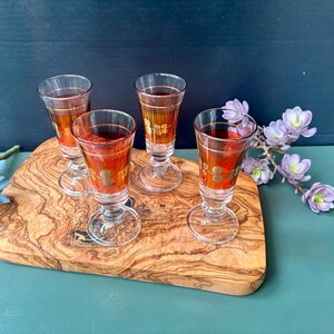 May include: Four small, clear glass shot glasses with gold accents and a red liquid inside. The glasses are arranged on a wooden cutting board with a light brown background.