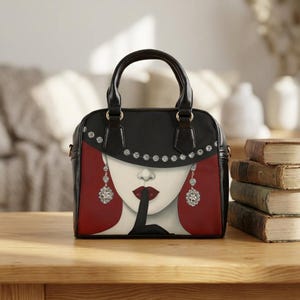 May include: A black handbag featuring a graphic illustration of a face wearing a wide-brimmed hat with silver studs, dangling crystal earrings, and red lipstick, with a finger held to the lips in a shushing gesture against a deep red background.