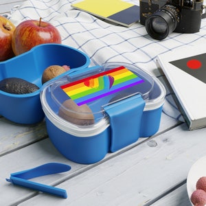 May include: A blue lunchbox with a rainbow heart and a rainbow flag design. The lunchbox has a clear lid and a removable latch. The lunchbox is sitting on a white and blue checkered tablecloth.