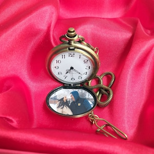 Può includere: Un orologio da tasca dorato con una foto di una coppia all'interno. L'orologio ha una catena e una chiusura.