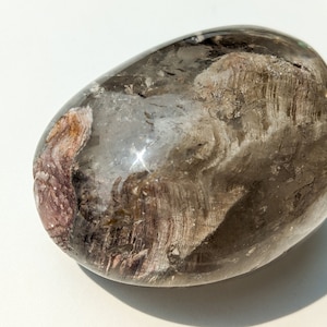 May include: A smooth, oval-shaped, smoky quartz crystal with brown and pink inclusions. The crystal is translucent and has a light brown color.