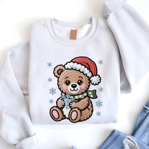 May include: White sweatshirt featuring a cartoon teddy bear wearing a Santa hat and a green scarf. The bear is holding a snowflake and surrounded by snowflakes. The design is embroidered. The sweatshirt is paired with blue jeans.