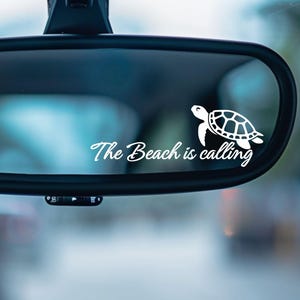 May include: A white vinyl decal applied to a car rearview mirror featuring a graphic of a sea turtle and the text The Beach is calling in a cursive script font. The decal is positioned on the glass surface of the black rearview mirror.