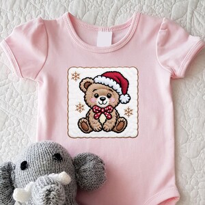 May include: A light pink baby onesie featuring a Christmas-themed teddy bear design. The bear wears a Santa hat and a red bow tie. The onesie has short sleeves and a snap closure. A gray elephant plush toy is in the foreground.