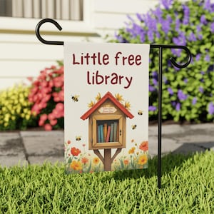 May include: A white garden flag with the text "Little Free Library" in red and a small red and brown birdhouse with books inside. The birdhouse has a sign that says "Take One Leave One". The flag is surrounded by yellow and orange flowers and bees.