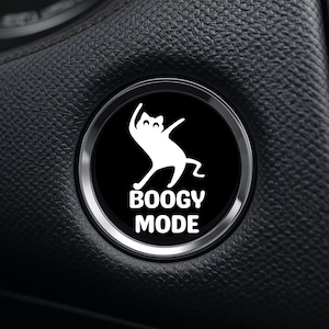May include: A black and silver car start button with the words "BOOGY MODE" in white text. A white cat silhouette is dancing above the text. The button is set in a black, textured surface.