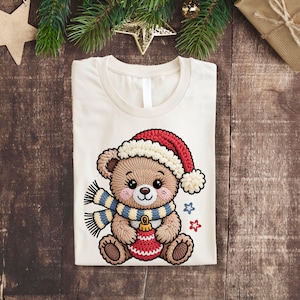 May include: Cream-colored t-shirt featuring a detailed embroidered teddy bear wearing a red and white Santa hat and a blue and yellow striped scarf. The bear is holding a red ornament. Two blue and red stars are also embroidered on the shirt.