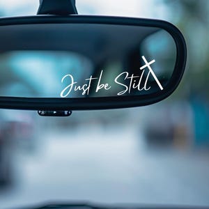 May include: A car's rearview mirror with the words "Just be Still" in white script, accompanied by a white cross. The mirror reflects a blurred outdoor scene with a soft, out-of-focus background.