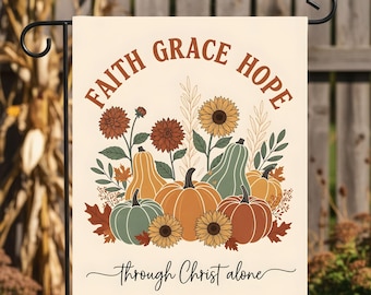 Puede incluir: Bandera de jardín color crema con las palabras "Faith Grace Hope" sobre una colección de calabazas, girasoles y follaje. La frase "through Christ alone" está escrita debajo. La bandera se muestra al aire libre.