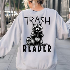 May include: Light gray sweatshirt with a black and white illustration of a raccoon reading a book. The text "TRASH READER" is printed above and below the raccoon graphic. The sweatshirt is a casual style.
