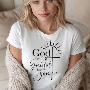 May include: A white short-sleeved t-shirt featuring black text that reads God i'm just Grateful for you, accompanied by a graphic of a cross with a sunburst design above it. The shirt is worn under a textured beige knit cardigan sweater.