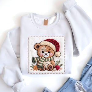 May include: White sweatshirt featuring an embroidered teddy bear wearing a red and cream Santa hat and a green and white striped scarf. The bear is surrounded by decorative elements, including small yarn balls and sprigs of greenery.