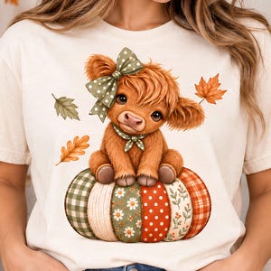 Könnte beinhalten: Ein cremefarbenes T-Shirt mit einer Grafik eines flauschigen braunen Hochlandrindes, das auf einem Patchwork-Kürbis sitzt. Das Rind trägt eine grüne Schleife und ein Halstuch mit Punkten. Der Kürbis hat Segmente mit Karos, Blumen und Punkten sowie Herbstblätter.