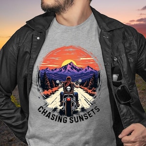 May include: Gray t-shirt with a graphic of a person riding a motorcycle towards a sunset over mountains. The text "CHASING SUNSETS" is below the graphic. The person is wearing a red bandana and a skull is on the back of the jacket.