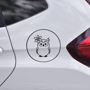 May include: A black and white decal featuring a cartoon owl with a flower on its head. The owl is inside a white circle, and the decal is affixed to a white car. The design is simple and clean.